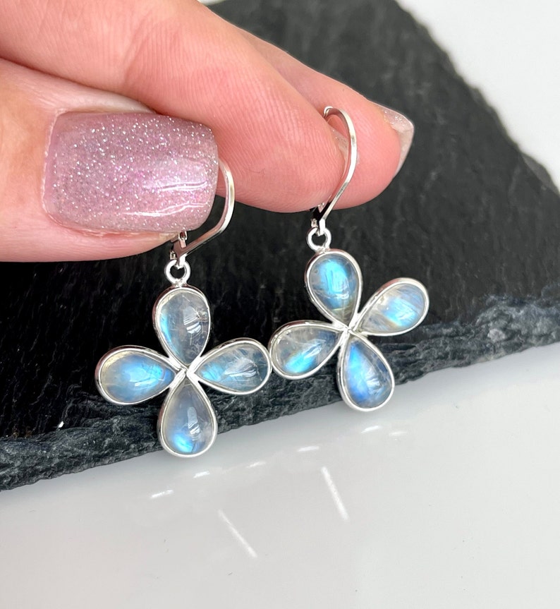 May include: A pair of silver-toned earrings featuring a four-petal flower design. Each petal holds a pear-shaped, iridescent, blue-white stone. The earrings are held up by a person's fingers against a dark background.