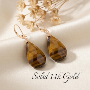 May include: A pair of drop earrings featuring faceted, teardrop-shaped tiger's eye gemstones suspended from solid 14k gold leverback hooks. The stones display characteristic brown and golden bands. The text Solid 14k Gold is visible below.