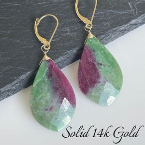 May include: A pair of teardrop earrings with gold-coloured clasps. The earrings feature green and maroon-coloured stones. The text "Solid 14k Gold" is written in cursive at the bottom of the image.