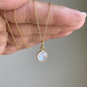May include: A gold-colored necklace featuring a small, round pendant with a light blue and white stone. The pendant is set in a gold bezel and hangs from a delicate gold chain. The necklace is displayed against a neutral background.