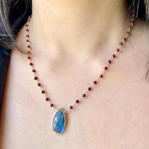 May include: A gold chain necklace with a teardrop-shaped blue labradorite gemstone pendant. The chain is made of small red beads.
