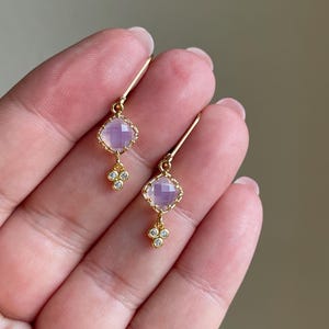 May include: A pair of gold earrings with a light purple square gemstone and a cluster of three small clear gemstones dangling below.