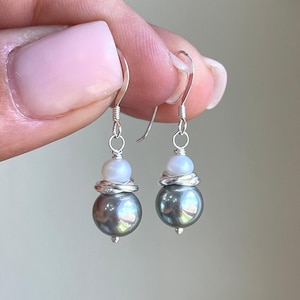 May include: A pair of dangle earrings featuring a large, dark gray pearl at the bottom, topped with a smaller white pearl and wrapped with silver wire. The earrings have silver fish hook ear wires.