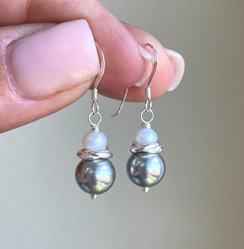 May include: A pair of dangle earrings featuring a large, dark gray pearl at the bottom, topped with a smaller white pearl and wrapped with silver wire. The earrings have silver fish hook ear wires.