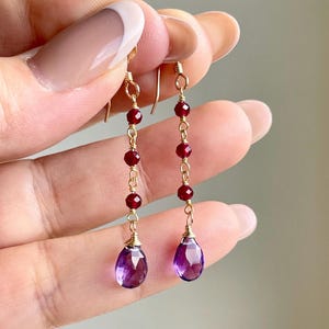 May include: Gold-toned dangle earrings featuring a series of small, round, red beads and a teardrop-shaped, faceted purple amethyst. The earrings are suspended from gold hooks.