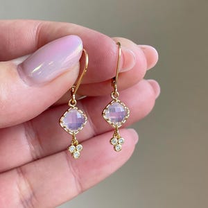 May include: A pair of gold earrings with a light pink square gemstone and a cluster of smaller clear gemstones dangling below.