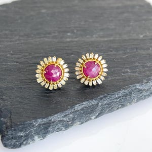 May include: A pair of stud earrings with a central, faceted, deep pink gemstone surrounded by a halo of smaller, rectangular, gold-coloured accents. The earrings are set in a gold-coloured metal.