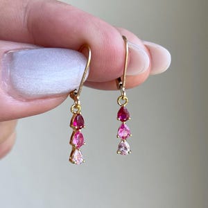 May include: Gold-tone drop earrings with three teardrop-shaped gemstones in shades of pink and red. The gemstones graduate in color from dark red to light pink. The earrings are held up by a gold hook.