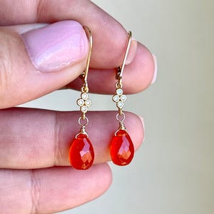 May include: Gold-tone drop earrings with a four-leaf clover design and teardrop-shaped, faceted orange gemstones. The earrings are held up by a person's hand. The earrings are a jewelry item.