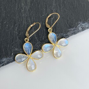 May include: A pair of gold-colored earrings with a four-petal flower design. Each petal is a light blue and white stone, set in a gold-colored frame. The earrings have a lever-back closure.