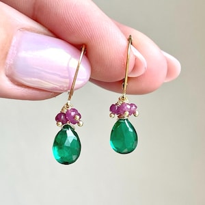 May include: A pair of gold-colored drop earrings. Each earring features a cluster of small, round, pink gemstones above a large, teardrop-shaped, emerald-green gemstone. The earrings are held up by a gold-colored hook.