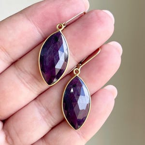May include: A pair of gold-toned earrings featuring marquise-shaped, faceted, deep purple gemstones. The gemstones are set in a gold bezel and have a hook closure. The earrings are designed for pierced ears and are a jewelry accessory.