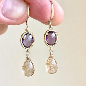 May include: Gold earrings with a purple oval gemstone and a teardrop-shaped brown gemstone dangling below.