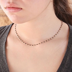 May include: A delicate gold chain necklace featuring small, evenly spaced black beads. The necklace rests against the neck, showcasing a minimalist design with a thin metallic link chain and dark, circular accents distributed along the entire length.
