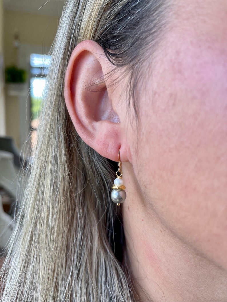May include: Gold-colored drop earrings with a gray pearl and a white pearl accent. The earrings have a gold wire design and a hook closure. The earrings are shown on a person's ear.