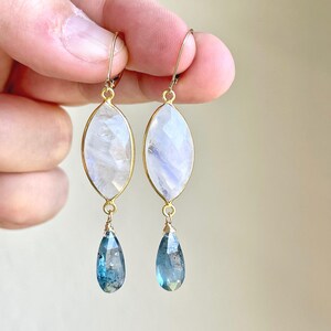 May include: A pair of gold-toned earrings. Each earring features a large, oval-shaped, faceted, white gemstone with a gold border, and a teardrop-shaped, blue gemstone dangling below. The earrings are held up by a person's fingers.