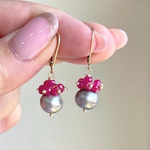 May include: A pair of gold-colored drop earrings. Each earring features a large, gray pearl at the base, topped with a cluster of small, red gemstones. The earrings are held by a gold-colored hook.