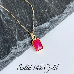 May include: A delicate gold chain necklace with a rectangular pendant featuring a vibrant pink gemstone. The pendant is set in solid 14k gold. "Solid 14k Gold" is written in cursive at the bottom of the image.