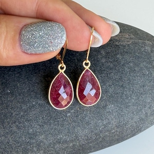 May include: A pair of gold-colored drop earrings with teardrop-shaped, faceted, deep red gemstones. The earrings are held up against a dark gray stone surface. The gemstones have a polished, reflective surface.