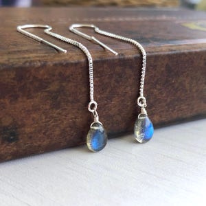 May include: A pair of silver threader earrings featuring delicate box chains and teardrop-shaped labradorite gemstones. Each stone displays a distinct blue flash, suspended from the end of the thin, polished silver chain links.