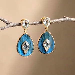 May include: A pair of gold drop earrings featuring large, pear-shaped blue labradorite stones with a textured surface. Each earring is accented with two clear, faceted pear-shaped stones and a small cluster of tiny white crystals at the base.