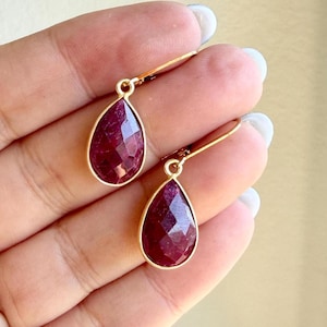 May include: A pair of drop earrings featuring faceted, teardrop-shaped deep red gemstones set in gold-toned metal frames. Each earring hangs from a simple gold-toned hook, showcasing a polished, reflective surface on the dark red stone facets.