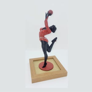Può includere: Statuetta di una ballerina in posa dinamica, con una gamba tesa e le braccia verso l'alto. La ballerina indossa un top rosso con cintura gioiello, leggings neri e scarpe nere. La statuetta è montata su una cornice di legno.