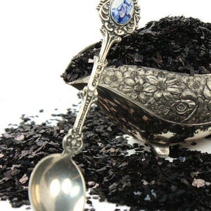 May include: A silver spoon with a blue and white floral design on the handle is filled with black glitter. The glitter is scattered around the spoon on a white surface.