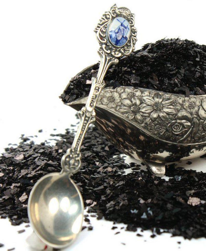 May include: A silver spoon with a blue and white floral design on the handle is filled with black glitter. The glitter is scattered around the spoon on a white surface.