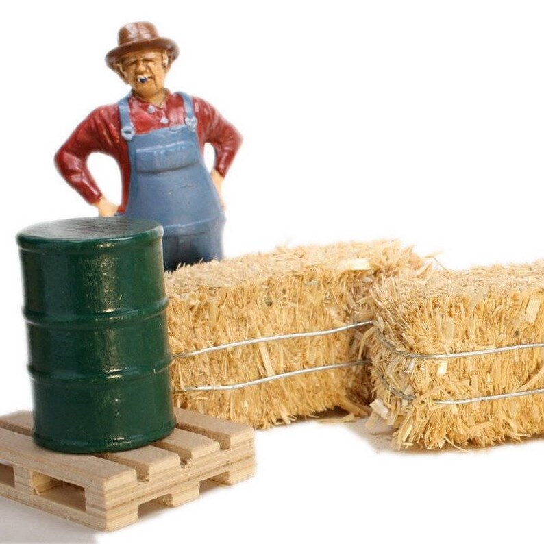 May include: A miniature figurine of a farmer wearing a straw hat and overalls stands next to a green barrel on a wooden pallet. Two bales of hay are in the background.