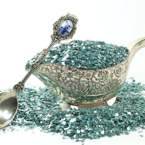 May include: A silver serving dish filled with teal blue iridescent glitter. The dish is decorated with a floral pattern. A silver spoon with a blue and white design is resting on the edge of the dish.