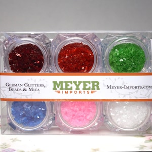 Pode incluir: Conjunto de seis pequenos frascos cheios de purpurina de diferentes cores. Os frascos estão dispostos em duas filas de três. Os frascos são etiquetados com "German Glitters, Beads & Mica" e "Meyer Imports".