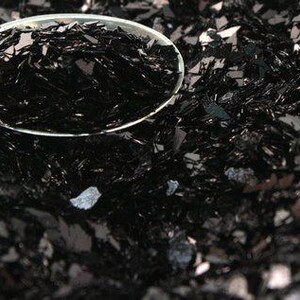May include: A close-up of black glitter, a popular material for crafting and decorating. The glitter is in a small spoon, showing its texture and sparkle.