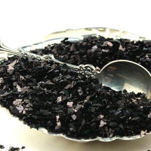 May include: A silver spoon scoops black iridescent glitter from a silver bowl. The glitter is a mix of small and large pieces, with a subtle shimmer.