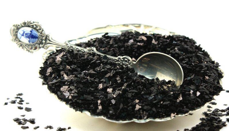 May include: A silver spoon scoops black iridescent glitter from a silver bowl. The glitter is a mix of small and large pieces, with a subtle shimmer.