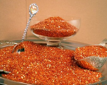 May include: A silver spoon scoops up orange glitter with gold flecks. The glitter is displayed in a glass bowl and a silver serving spoon.