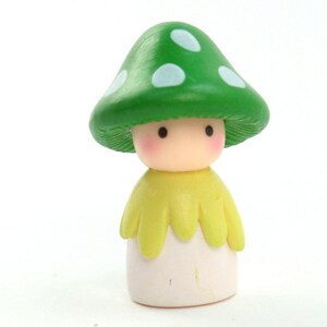 May include: A small green mushroom figurine with white spots, wearing a yellow cape and a white dress. The figurine has a cute, cartoon-like face with black eyes and pink cheeks.