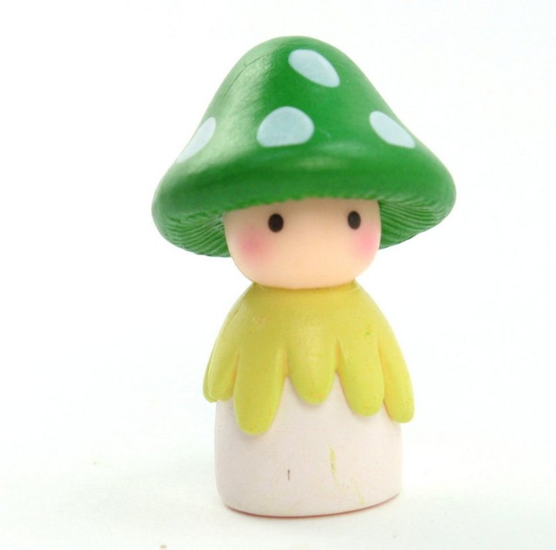 May include: A small green mushroom figurine with white spots, wearing a yellow cape and a white dress. The figurine has a cute, cartoon-like face with black eyes and pink cheeks.