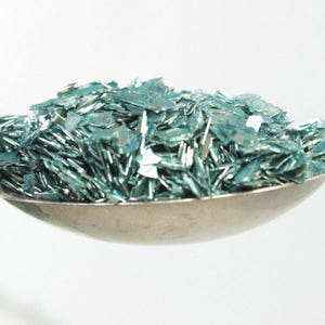 May include: A silver spoon filled with teal blue glitter. The glitter is made of small, flat pieces that reflect light.