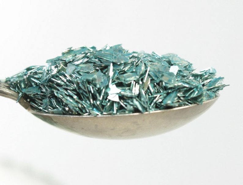 May include: A silver spoon filled with teal blue glitter. The glitter is made of small, flat pieces that reflect light.