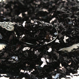 May include: Black iridescent glitter flakes in a spoon. The glitter is a mix of small and large pieces, with a metallic sheen.