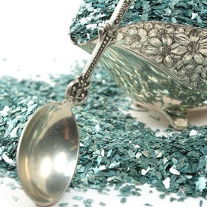 May include: A silver serving spoon filled with teal blue iridescent glitter. The glitter is scattered around the spoon on a white background.