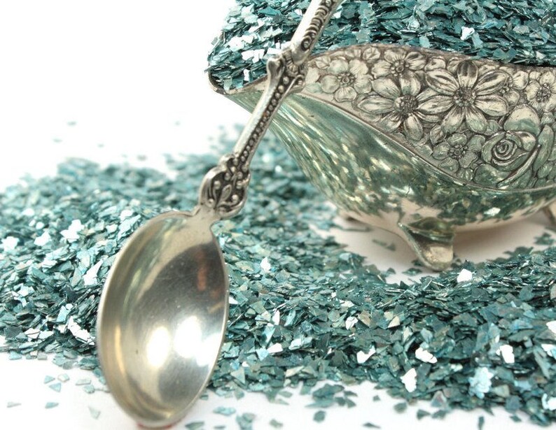May include: A silver serving spoon filled with teal blue iridescent glitter. The glitter is scattered around the spoon on a white background.