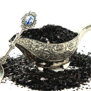 May include: A silver serving dish with an ornate floral design, filled with black flakes. A silver spoon with a blue and white porcelain handle rests on the edge of the dish.