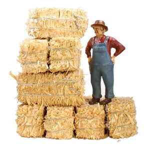 May include: A miniature figurine of a farmer wearing a straw hat and overalls stands in front of a stack of hay bales. The hay bales are tied with twine and are a light brown color.