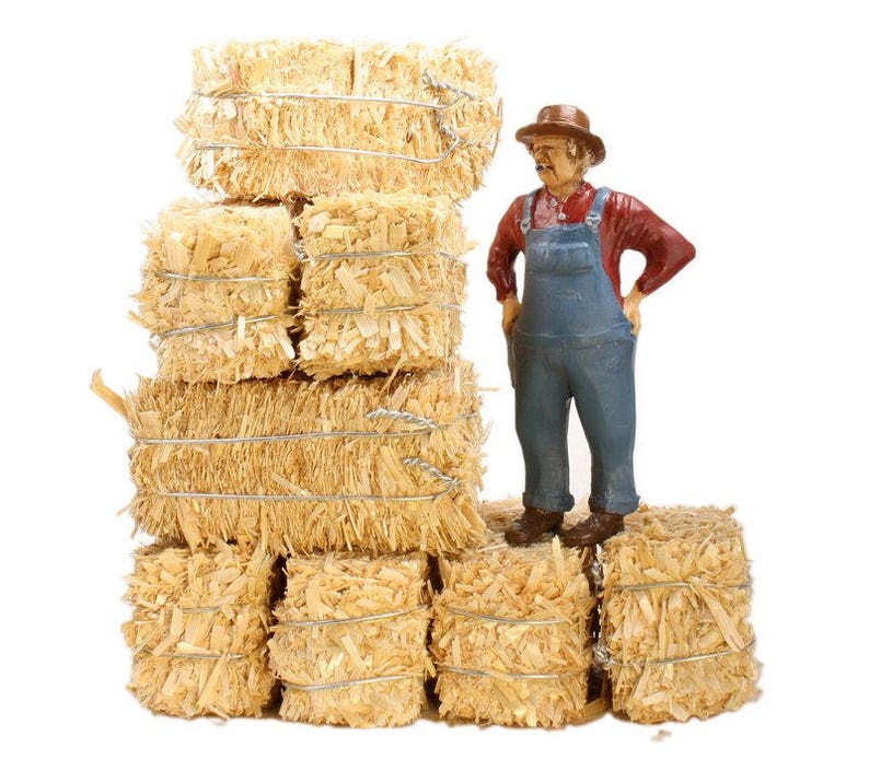 May include: A miniature figurine of a farmer wearing a straw hat and overalls stands in front of a stack of hay bales. The hay bales are tied with twine and are a light brown color.