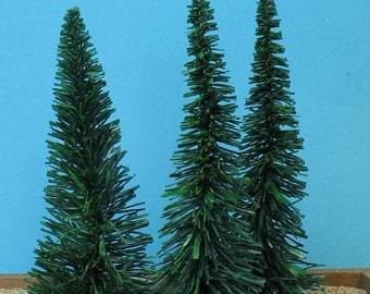May include: Three miniature artificial Christmas trees with green needles. The trees are of varying heights and are set on a bed of sand. The background is a solid light blue.