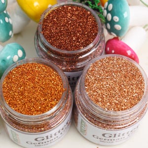 Może przedstawiać: Trzy małe szklane słoiki wypełnione różnymi odcieniami miedzianego brokatu. Słoiki mają białe etykiety z nadrukiem "Glitter German Fine Glitter".