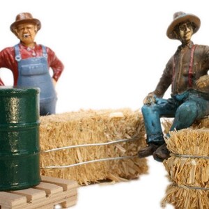 May include: Two miniature figurines of farmers, one wearing a red shirt and blue overalls, the other wearing a brown jacket and blue jeans, sitting on bales of hay. A green barrel sits on a wooden pallet in front of the figurines.