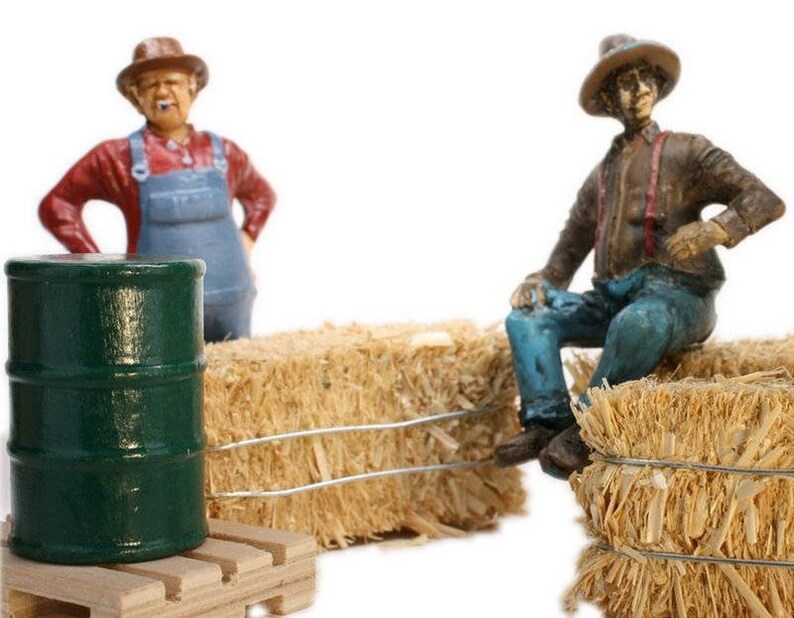 May include: Two miniature figurines of farmers, one wearing a red shirt and blue overalls, the other wearing a brown jacket and blue jeans, sitting on bales of hay. A green barrel sits on a wooden pallet in front of the figurines.